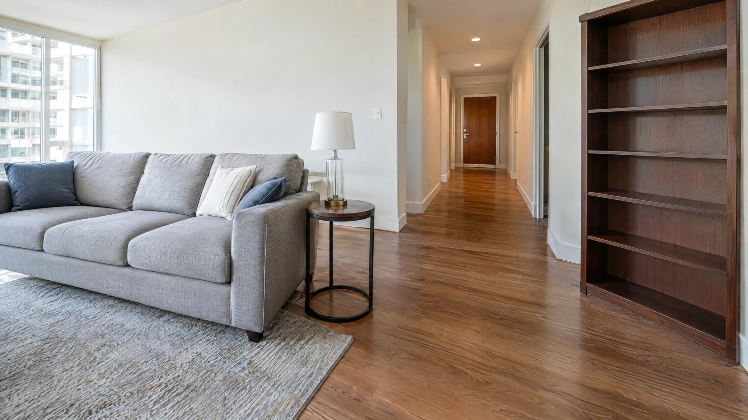 A sofa, side table and empty bookcase inside a vacant condo with a clear hallway to the door.